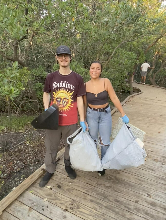 Two volunteers on a wooden boardwalk smile while holding large bags of collected trash after a coastal cleanup.