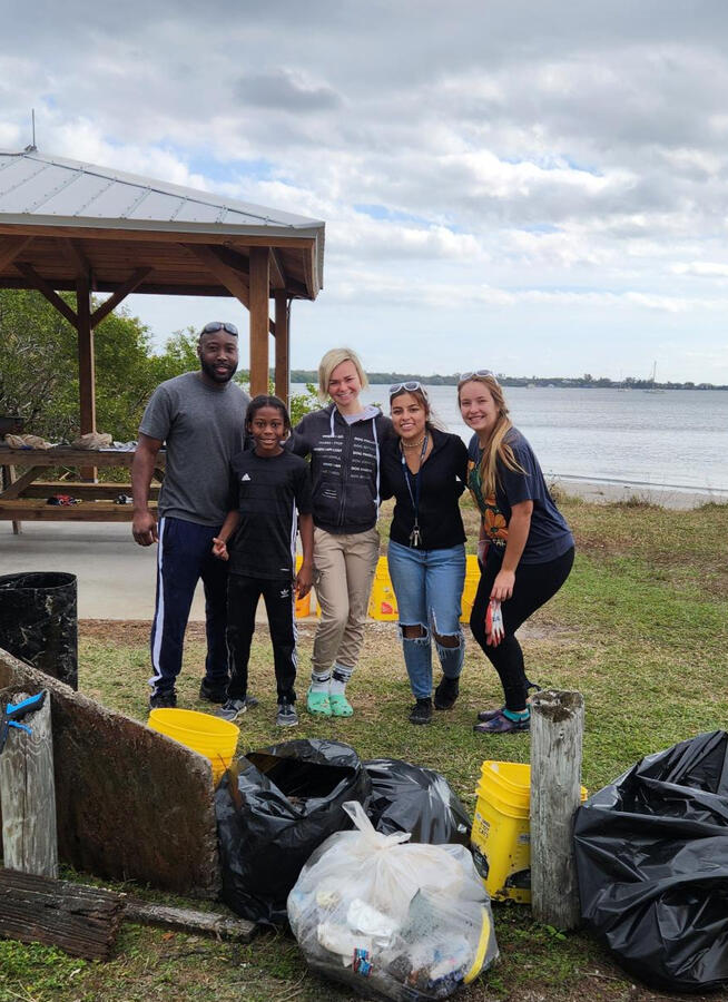 A group of five volunteers pose together by the water with full trash bags and yellow buckets, marking the completion of their cleanup effort.