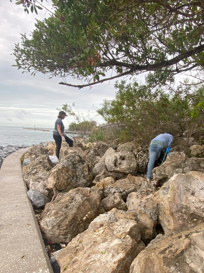 City Island Two volunteers pick up trash among large rocks along the shoreline during a coastal cleanup.