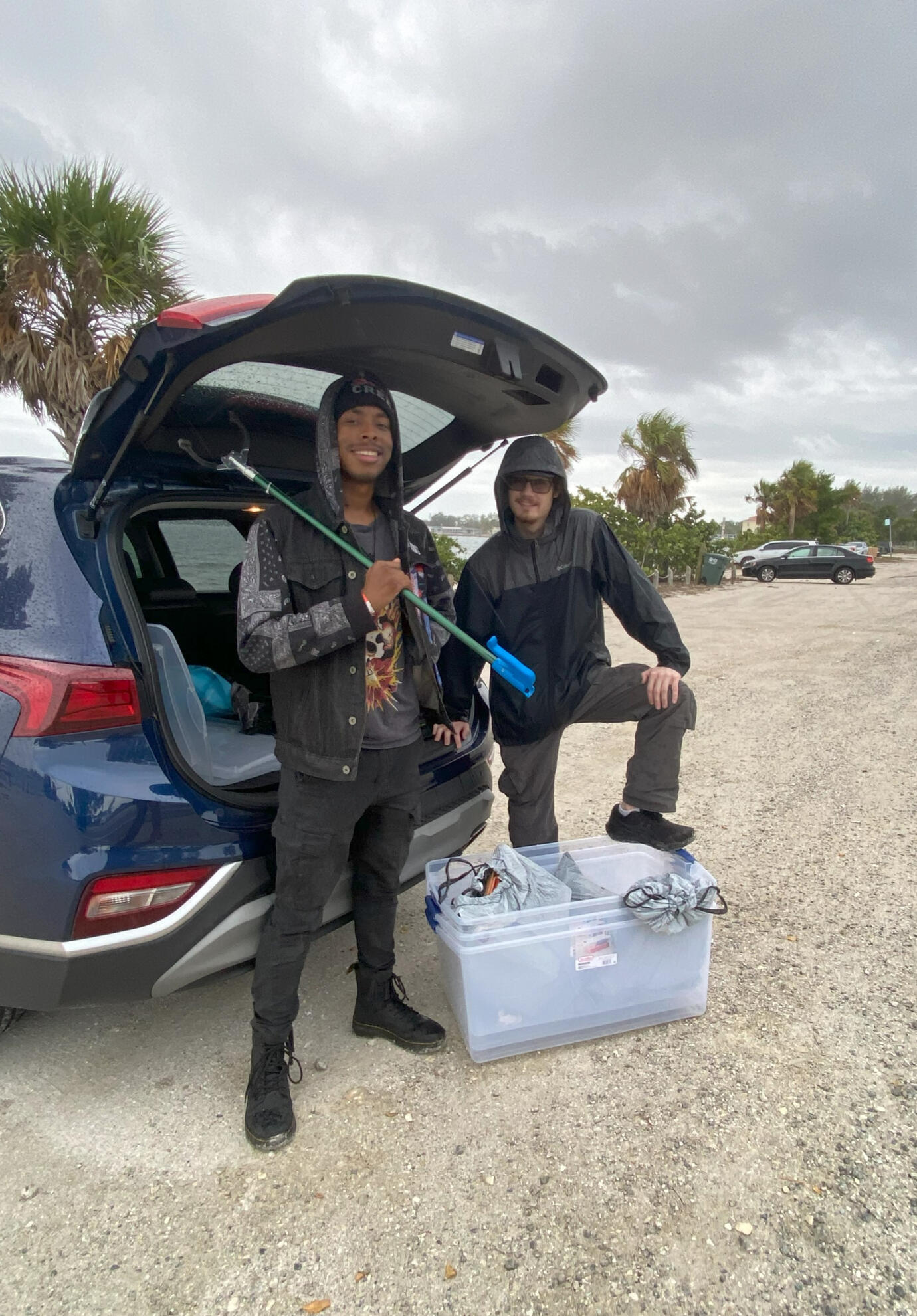 Palma Sola Cleanup Two volunteers stand beside a car with cleanup tools and supply bins, preparing to wrap up after a coastal trash cleanup on a cloudy day.