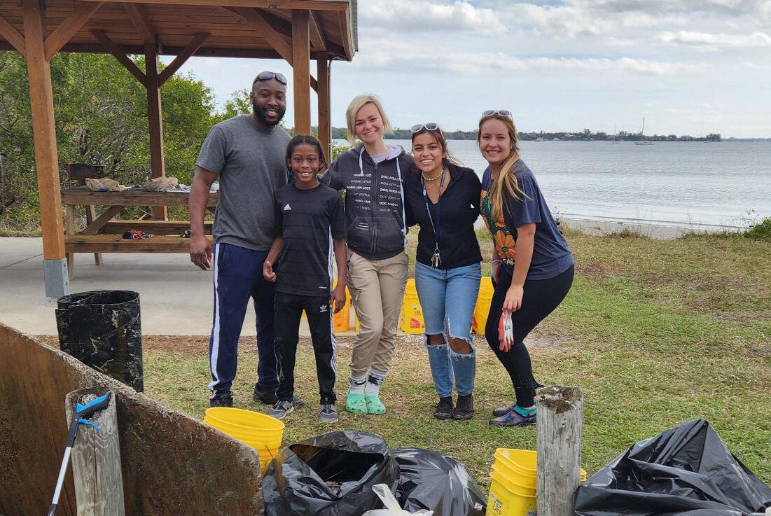 A group of five volunteers pose together by the water with collected trash bags and buckets, celebrating the end of a coastal cleanup.