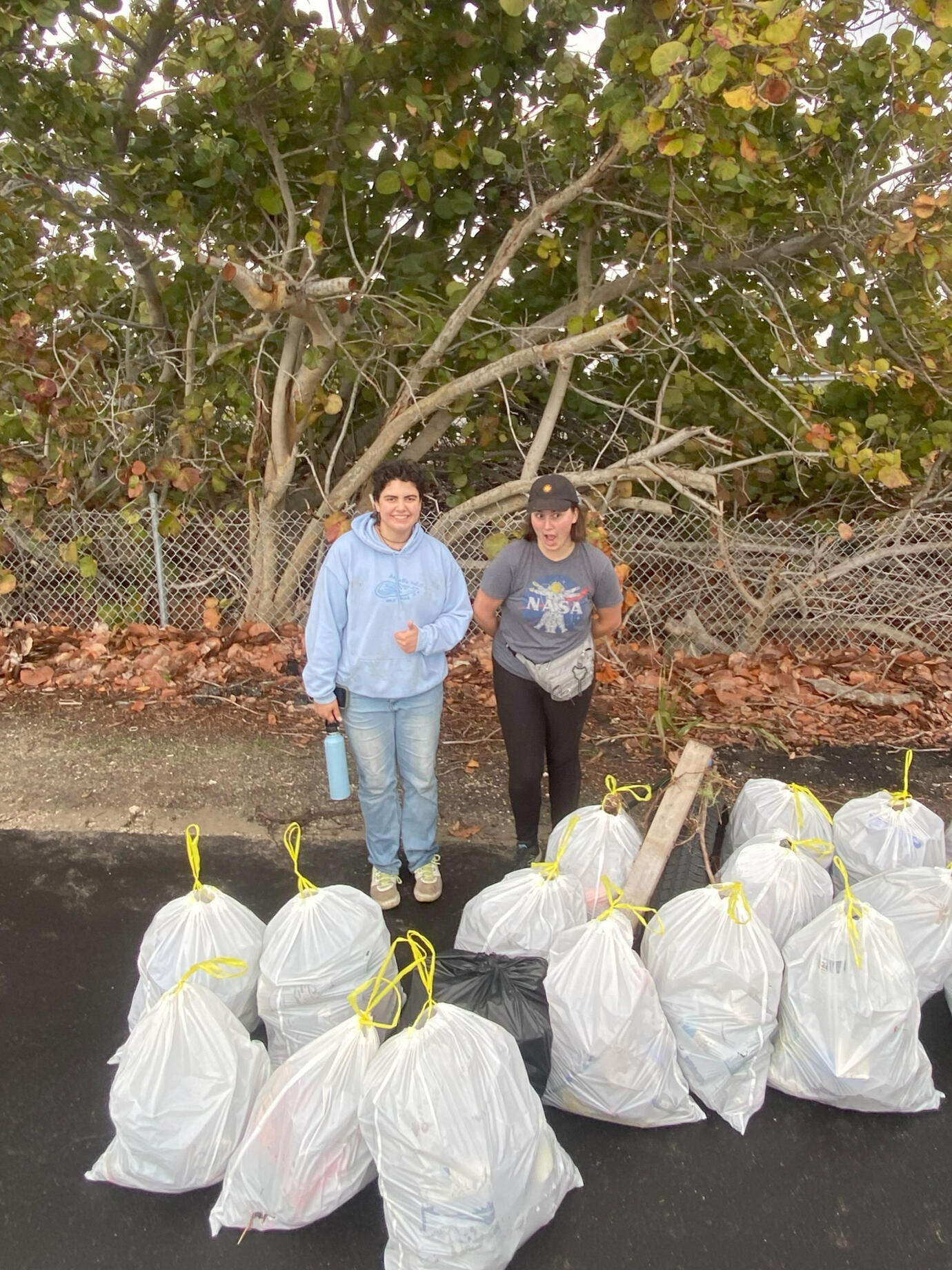 Two volunteers stand beside multiple filled trash bags after finishing a shoreline cleanup near coastal vegetation.