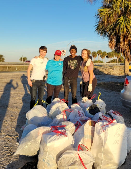 Four volunteers smile behind a large pile of filled trash bags, marking the completion of a successful beach cleanup.